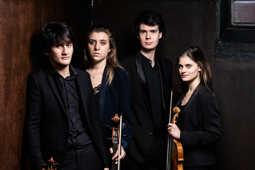 The four artists stand in front of a dark background and, holding their instruments, look at the camera.