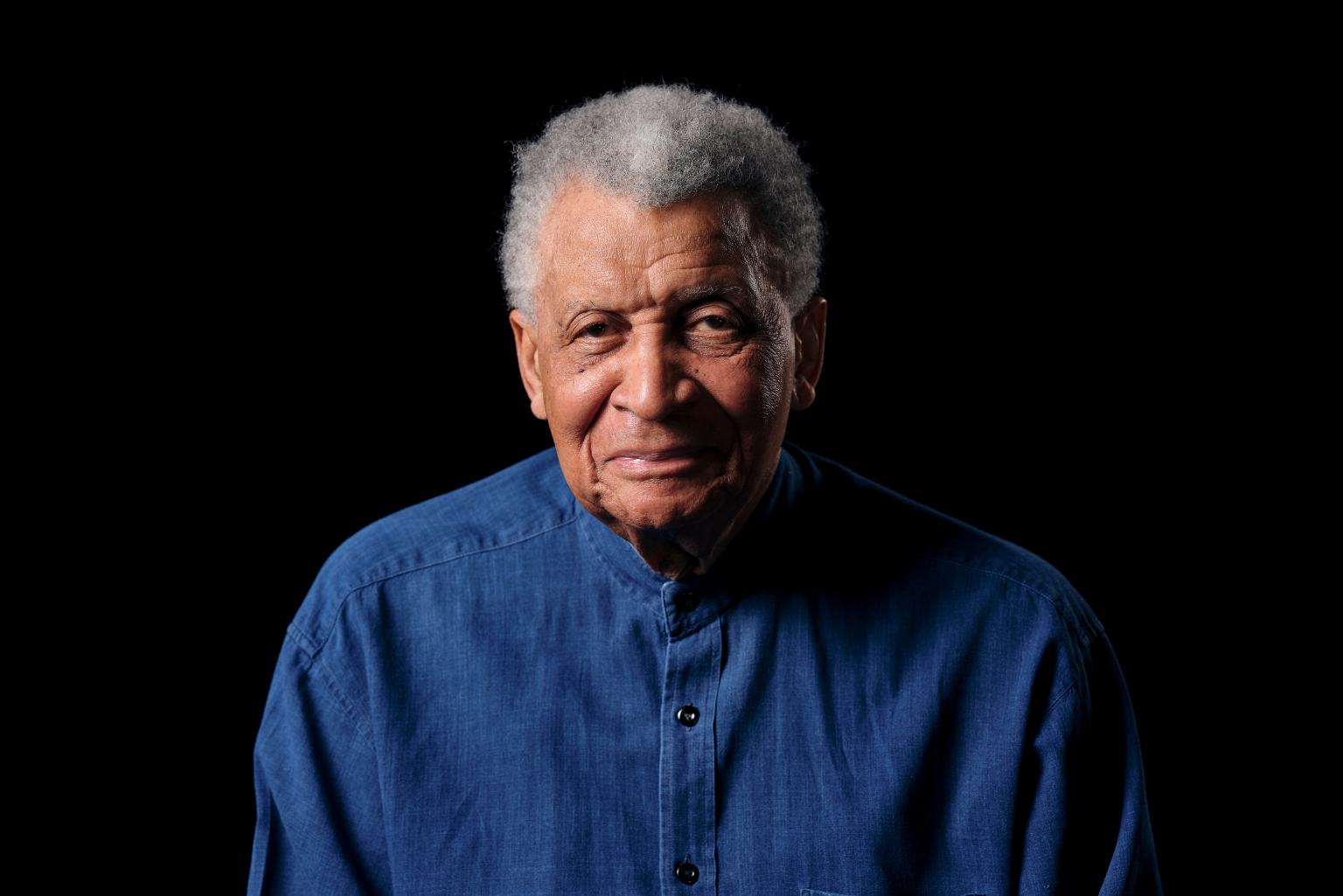 Abdullah Ibrahim sits with a black background and faces the camera.